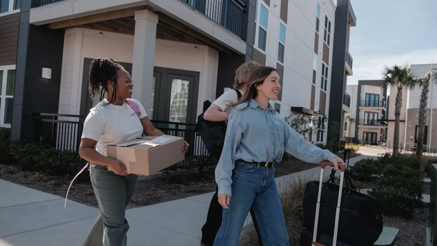 Two students carrying boxes and luggage into an apartment complex